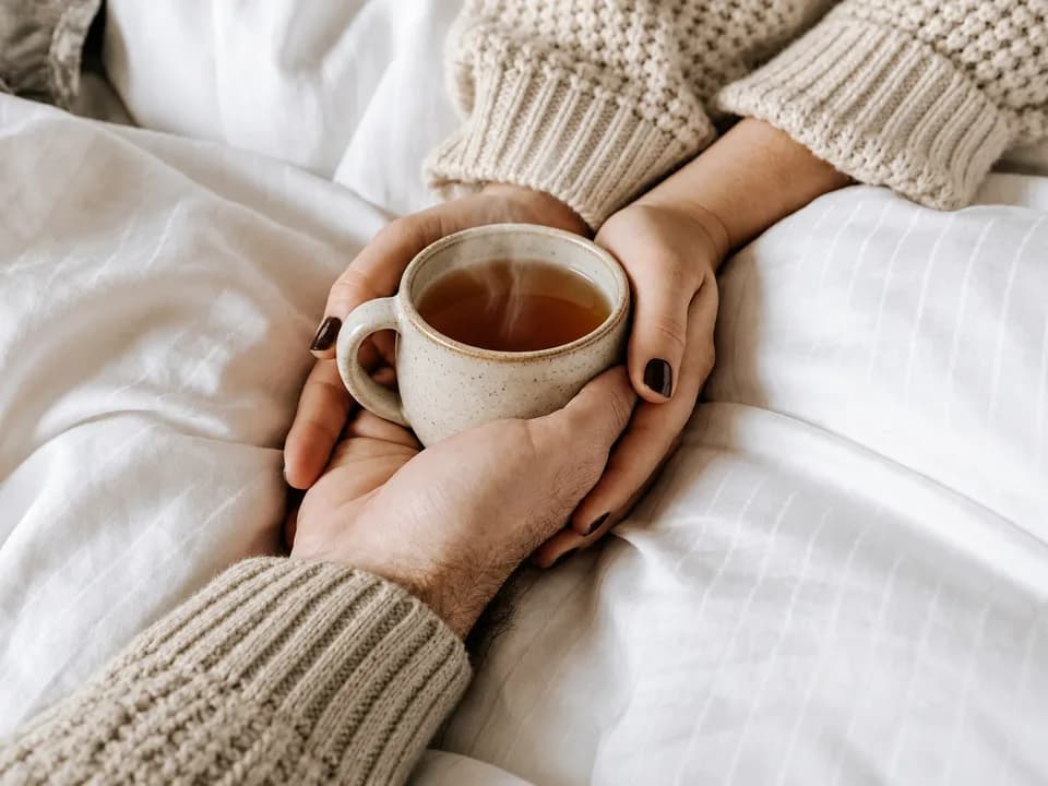 Hands around a warm mug of tea on soft white bedding