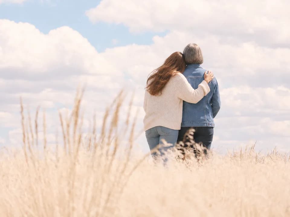 A daughter with her arm around her mother as they look out over a field of tall grass