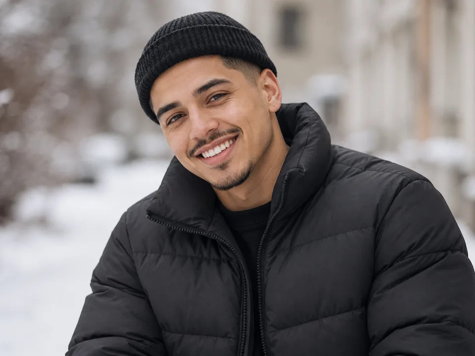 A young man in a beanie and puffer jacket smiling on a snowy street