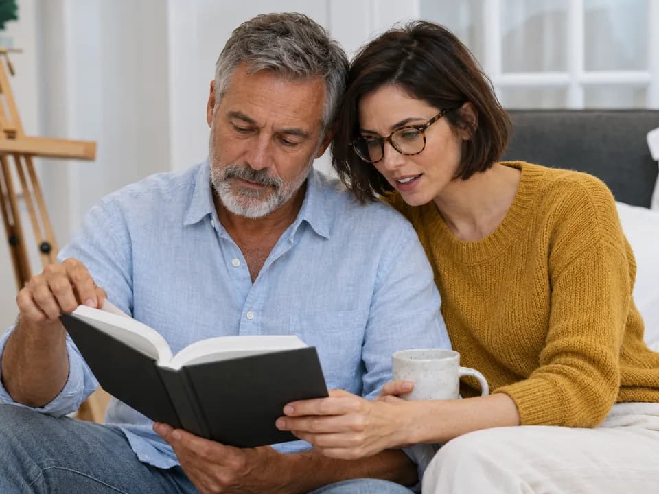 A grey-bearded man reading a book while a woman in glasses reads over his shoulder